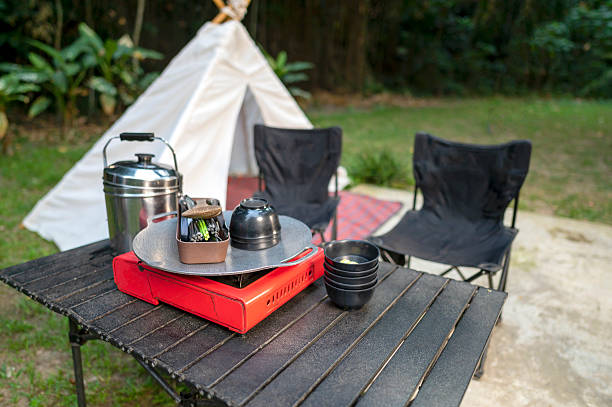 istock Portable camping table is set up outdoors with a cooking stove, pot, and chairs, with a white tent in the background. A scene of outdoor camping and cooking. 2231993052