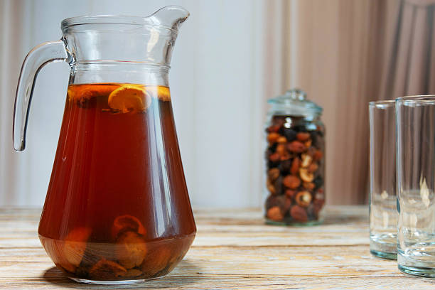 Glass pitcher of homemade dried fruit compote with lemon slices on rustic wooden table. Jar of dried fruits and empty glasses in cozy kitchen setting