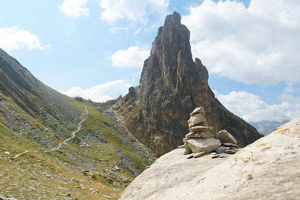 A majestic rock formation rises against a partly cloudy sky, showcasing the beauty of the natural landscape. In the foreground, a carefully balanced stack of rocks, known as a cairn, rests on a large boulder, marking the trail. A winding hiking path leads towards a group of people in the distance, suggesting a shared adventure and connection with nature. This scene evokes a sense of peacefulness and the spirit of exploration.