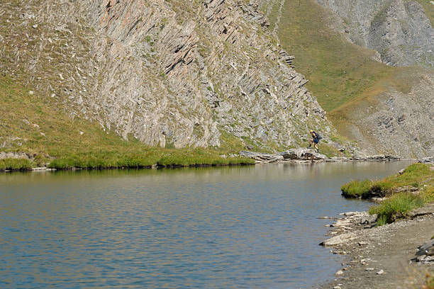 A hiker with a backpack and their dog traverse rocky terrain alongside a serene alpine lake, surrounded by a rugged mountain landscape. The scene evokes a sense of adventure and the beauty of nature in a remote, peaceful setting.