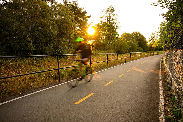 Motion-blurred cyclist rides into the golden sunrise along a bike path lined with trees and wildflowers.