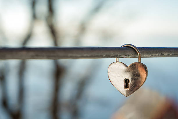 A small iron heart-shaped lock hangs on an iron rod against a blurred background The surface of the lock is slightly scratched and sanded and is located on the right side of the picture.