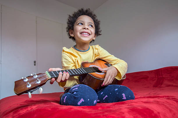 istock Happy child playing ukulele on a cozy red bed in a sunny room 2232046536