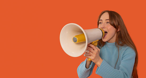 istock Young woman enthusiastically speaking into a megaphone against a bright orange background during a spirited rally or event 2232048273
