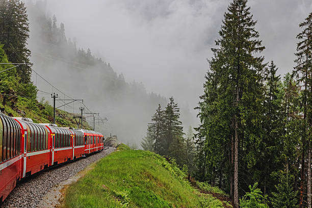 A red train winds along a narrow track amidst a misty mountain landscape in Switzerland