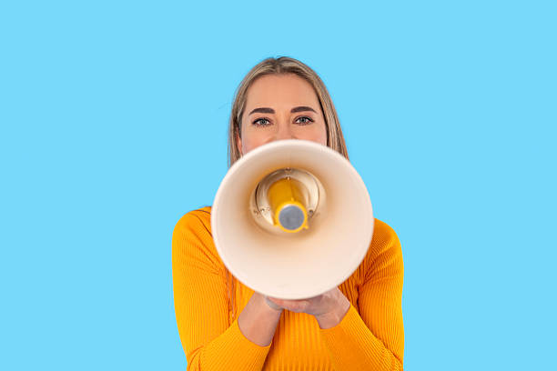 istock Young woman in bright yellow sweater holds megaphone against blue background while communicating a message with enthusiasm and energy 2232054253