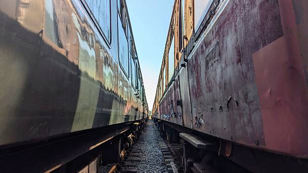 Between two train carriage cars, wheels, concrete sleepers, rusting railroad tracks. Rusty derelict undercarriage of obsolete public transport network railway line. Rail transportation commuting journey, railcar travelling straight to horizon vanishing point. Deserted dilapidated carriages, rails, sleeper on national rail transit. Travel or commute apocalyptic imagery of abandoned industrial railcar on train track.