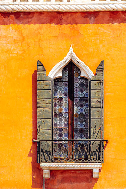 A traditional Venetian window framed with Gothic detailing and wooden shutters, set against a vibrant orange façade. The image showcases classic architectural charm and bright, inviting colors.