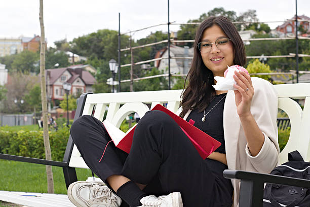 Young Indian college student sitting on a bench outdoors, reading a book and eating a croissant, symbolizing modern academic lifestyle, balance of study, leisure, and pursuit of knowledge.