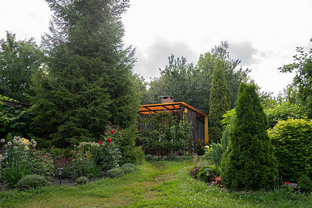 A picturesque garden path surrounded by dense greenery and flowers, leading to a small structure with an orange roof under a cloudy sky. Horizontal orientation.