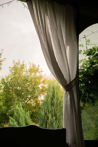 Soft white curtain blowing gently on a cozy garden terrace with trees and bushes visible through the window. Vertical frame capturing peaceful garden view.