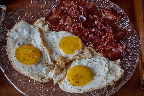 Hot scrambled eggs with bacon slices and two fresh croutons on a round white plate with cutlery on a light wooden table. Top view, flat lay.