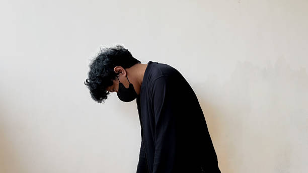istock A man in a black mask and dark outfit standing against a plain wall, looking down with a slouched posture, expressing tired 2232085817