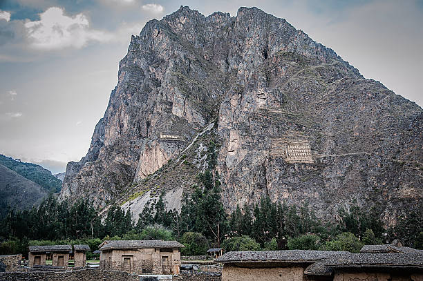 A towering, rugged mountain dominates the landscape behind a small Andean village, where traditional stone buildings with thatched roofs nestle among tall trees. The citadel of Pisac is built into the steep rock face and features two prominent rectangular structures—ancient ruins arranged in a grid-like pattern.