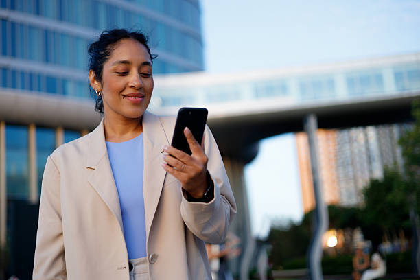 istock Professional woman working on smartphone, standing amid sleek skyscrapers with sunlight illuminating urban business environment 2232090427