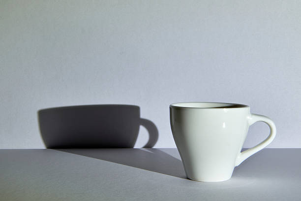 A single white mug casting a strong shadow on a grey background. A minimalist, high-contrast still life representing simplicity, duality, or the start of a new day.