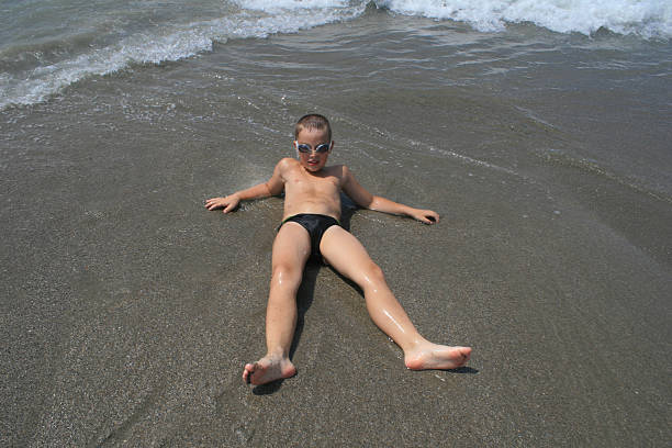 A boy from 6 to 9 years old in dark swimming trunks, swimming goggles lies on the sea sandy shore, sea waves, looks at the camera, view from above.