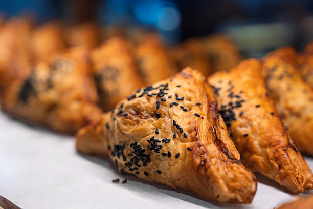Close-up of golden brown puff pastries topped with black sesame seeds, displayed in a bakery. Flaky texture and appetizing appearance highlight artisanal baking and indulgent snack culture.
