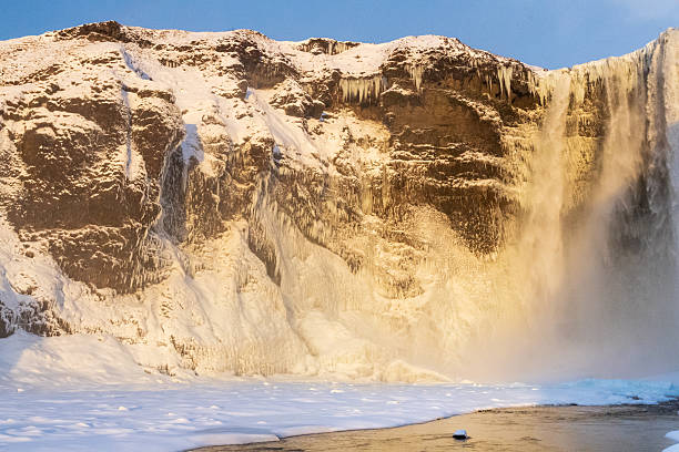 A bleak golden hour sun illuminates a partially frozen Skogafoss waterfall in southern Iceland. Skogafoss is one of the most spectacular and famous waterfalls in Iceland.