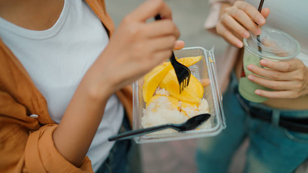 A close-up view of a young Asian woman having a taste of mango while going to a street food market with her friend.