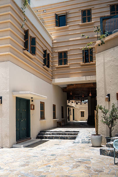 A sunlit courtyard in Al Muftaha Art Village is featuring traditional architecture with beige walls and wooden windows. Potted plants are adorning the stone pavement, creating a serene and inviting atmosphere.