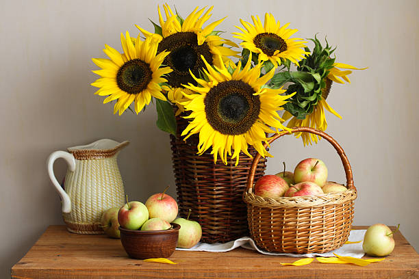 Sunflowers arranged in a wicker basket alongside ripe apples in a bowl and basket, forming a rustic still life composition on a wooden table