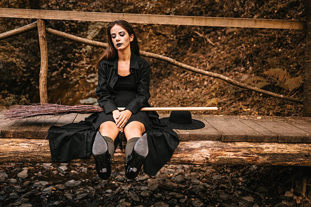 Young woman dressed as a witch sits sadly on a wooden bridge in the forest. She wears a long black dress, black coat, and dark makeup with black lipstick. Next to her are a broom and a witch hat, Halloween atmosphere with autumn colors in the background.