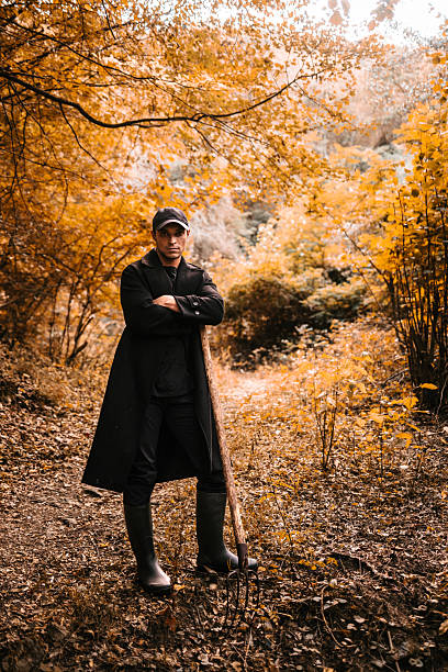 Serious man standing in the woods during autumn, dressed in a long dark coat and rubber boots, holding a pitchfork and looking into the distance. Halloween concep
