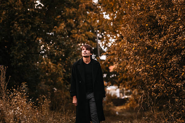 A man in a long black coat walking alone in an autumn forest, evoking a mysterious and eerie Halloween vibe. Halloween fashion model