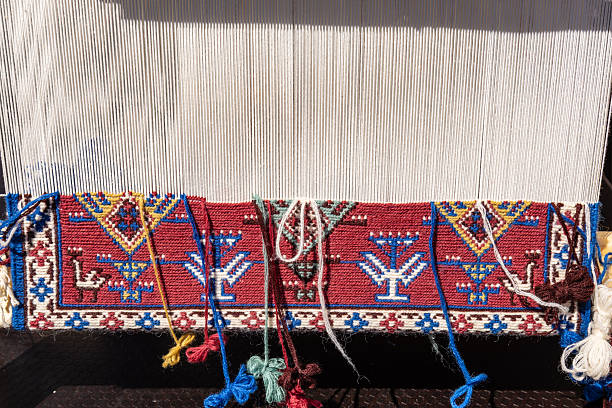 A close-up view of a traditional wooden loom with a partially woven carpet below. The vibrant red, blue, green, and yellow threads form an intricate geometric pattern, showcasing artisanal craftsmanship and cultural heritage in textile making.