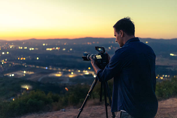 A photographer on a hill operates a professional camera on a tripod, capturing a serene cityscape under warm, golden hour light, with blurred urban lights sparkling in the distance.