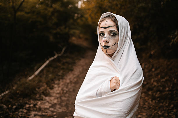Woman with horror makeup wrapped in white sheet standing in autumn forest