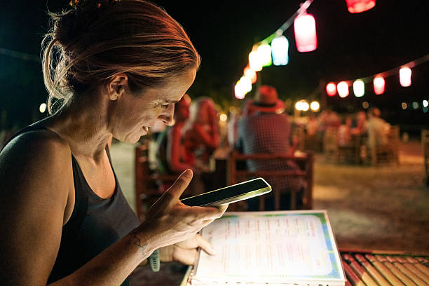 Woman reading menu with phone flashlight at night beach restaurant