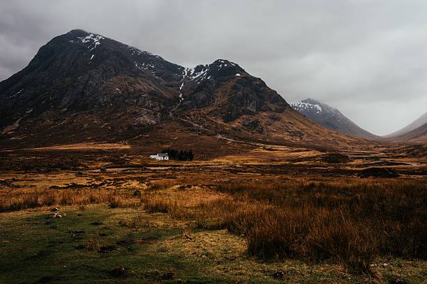 A remote white cottage nestled in the vast Scottish Highlands with towering mountains under a cloudy sky.