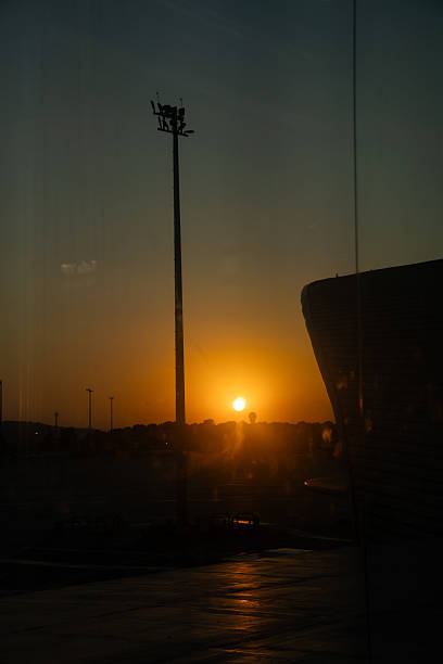 A vivid vertical photo of the setting sun reflected on a modern airport terminal window, with warm tones and strong silhouettes of poles and architectural lines.