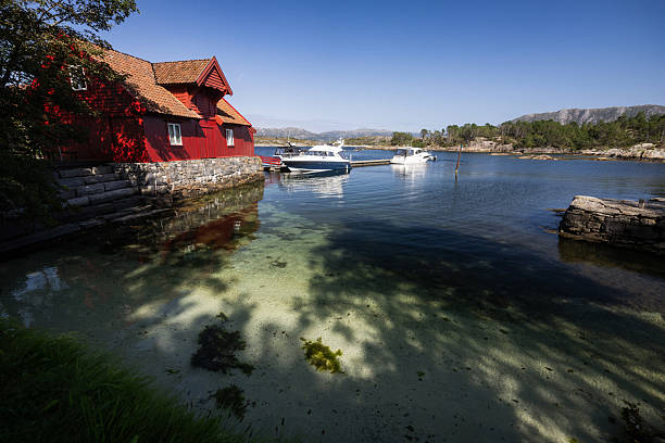 Scenic coastal view with motorboats moored near a pier beside a traditional red wooden boathouse, surrounded by clear blue water and rocky landscape in Norway.