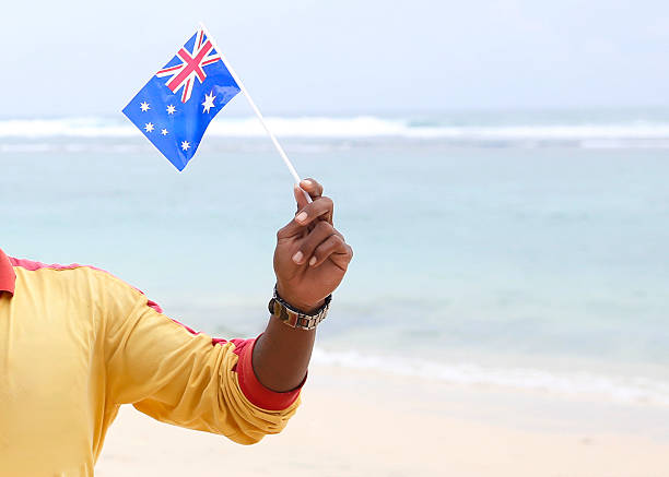 Close-up shot of a man in a yellow shirt holding a small Australian flag on a stick, standing on the beachside, ready for a national day event or patriotic celebration.\n\nConcept theme: Australia Day spirit, national flag in hand, patriotic pride, people celebrating independence day at the beach