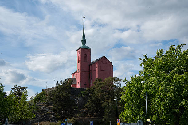 Nynashamn, Stockholm County, Sweden - May 26, 2025: A pink church with a green spire sits on a hill. The sky is cloudy and the sun is shining through the clouds
