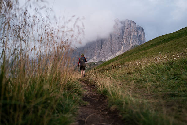 Traveler hiking on a mountain trail towards the dramatic cliffs of the Sella Group in the Dolomites, Italy. The rocky peaks rise into the clouds, creating a moody and atmospheric alpine landscape.