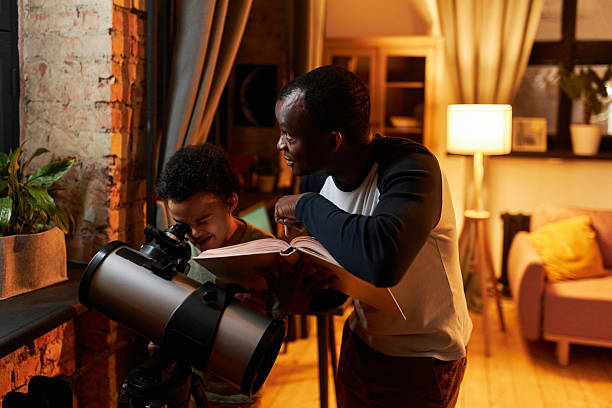 Mature African American father standing in front of telescope holding book telling his son about universe, when watching stars using telescope