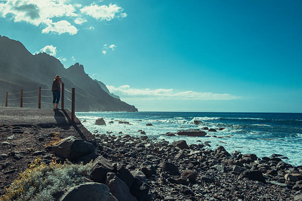 Traveler with hat walking on a seaside promenade overlooking the Atlantic Ocean in Tenerife, Canary Islands, Spain. The dramatic volcanic coastline, rocky shore, and bright sunlight create a scenic and relaxed atmosphere.
