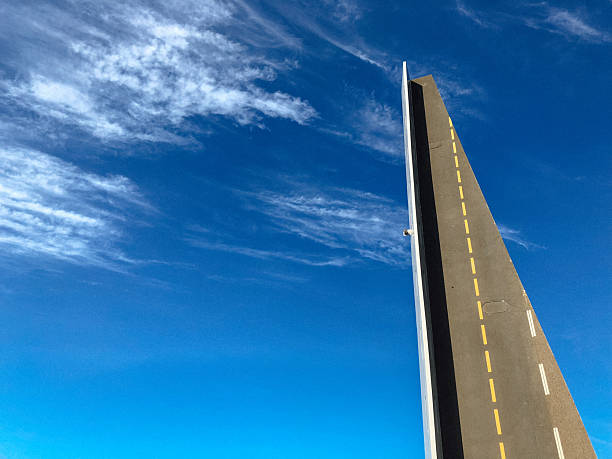 Dramatic perspective of a steep road bridge in Bergen, Norway, captured against a vivid blue sky with light clouds. The minimalist composition emphasizes the upward lines and creates a surreal impression of a road leading into the sky.