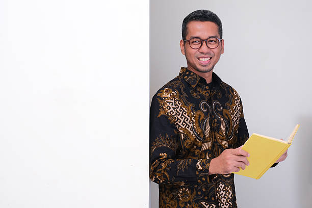 istock Indonesia school teacher standing beside blank wall holding a book with happy face expression 2232198112