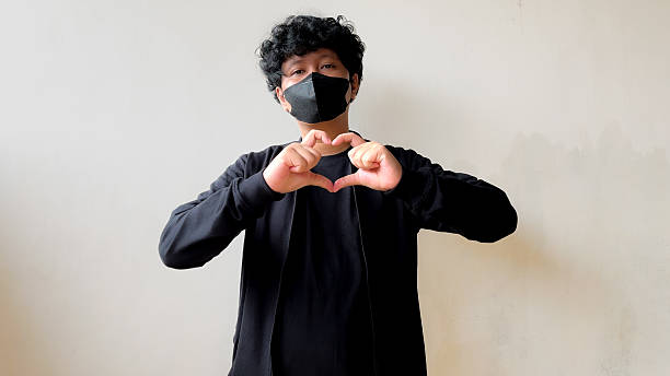 istock A man in a black outfit and mask standing against a plain wall, making a heart shape hand gesture using both hands by joining thumbs and index fingers. 2232207976