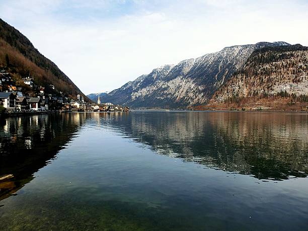 This breathtaking image captures a peaceful lakeside village nestled between majestic mountain ranges. The calm, mirror-like surface of the lake beautifully reflects the surrounding peaks and charming buildings along the shore, creating a harmonious and serene composition. Ideal for themes related to alpine travel, nature retreats, scenic landscapes, and tranquil living. Perfect for editorial use, travel promotions, wellness content, and environmental storytelling.