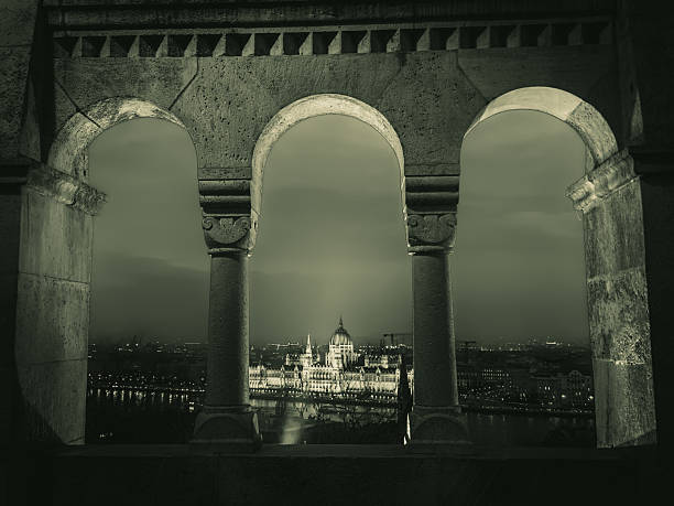 A married couple is standing by the Danube River with the Hungarian Parliament Building in the background during a spring weekend trip in Budapest, photographed on black and white 35mm analog film with an analog camera.