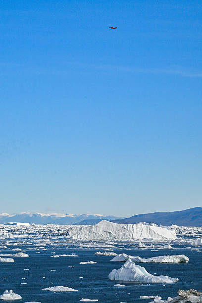 A small airplane flying above a seascape filled with icebergs and sea ice in Disko Bay, Greenland, with distant snow-covered mountains under a clear blue sky