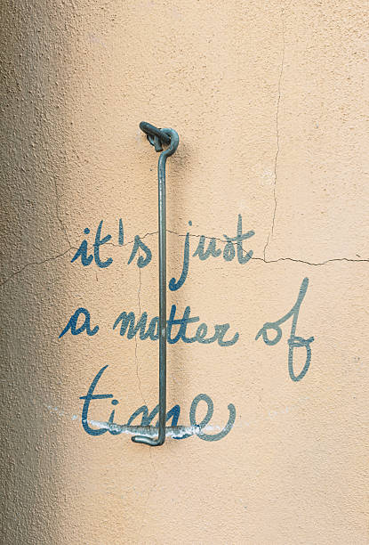 A long metal wall hook hangs vertically over a handwritten phrase reading “it’s just a matter of time,” painted in blue on a textured, cracked stucco wall. The hardware visually evokes a pendulum, while abrasions and weathering imply erosion and the passage of time. ***The text was digitally superimposed and a release is provided***