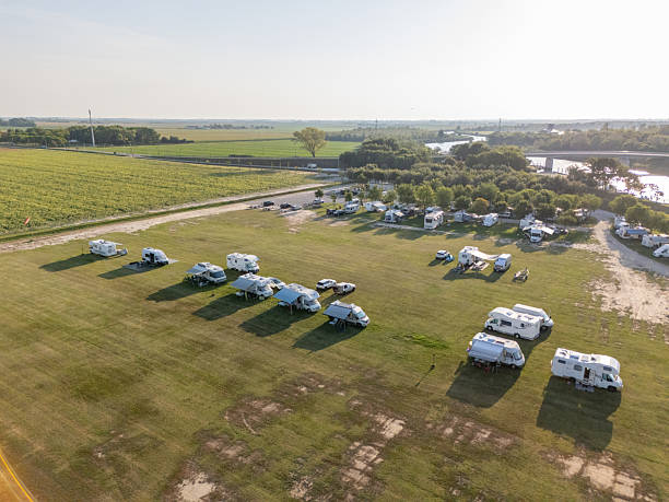Aerial photo of recreational vehicles parked in a large grassy field near trees and water. RV camping in rural area with open space and natural surroundings. Outdoor travel and vacation lifestyle.