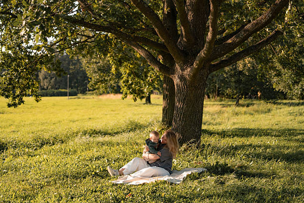 A mother and her baby are sitting on a blanket under a large, thick tree in a green summer park, creating an atmosphere of calm and care.